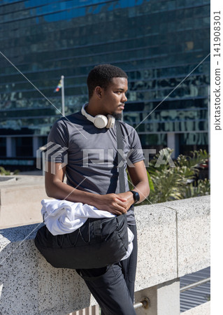 African American male leaning on railing at plaza, wearing grey tee, headphones, crossbody bag African American male leaning on railing at plaza, wearing grey tee, headphones, crossbody bag 141908301