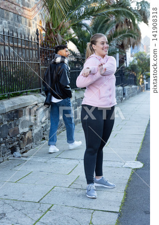 Diverse pedestrians on sidewalk with woman stretching in pink jacket, man carrying crossbody bag 141908318