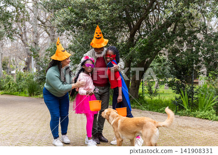 Family group standing on paved garden path wearing pumpkin hats, holding orange buckets with dog Family group standing on paved garden path wearing pumpkin hats, holding orange buckets with dog 141908331