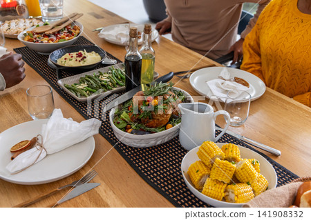 Diverse friends sitting at wooden table eating roast chicken with gravy jug and yellow sweater seen Diverse friends sitting at wooden table eating roast chicken with gravy jug and yellow sweater seen 141908332