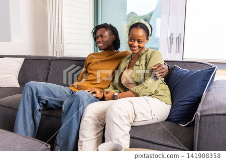 African American couple sitting on gray sectional couch in lounge holding coffee and navy pillow African American couple sitting on gray sectional couch in lounge holding coffee and navy pillow 141908358