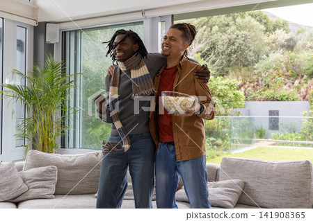 Diverse male friends standing close together in living room holding popcorn bowl celebrating 141908365