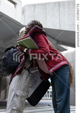 Diverse students hugging on campus under concrete overhang holding colorful notebooks, knit beanie Diverse students hugging on campus under concrete overhang holding colorful notebooks, knit beanie 141908778