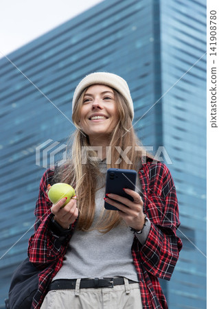 Woman in knit beanie red plaid shirt standing before glass tower with green apple and smartphone Woman in knit beanie red plaid shirt standing before glass tower with green apple and smartphone 141908780