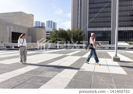 Two women crossing tiled urban plaza holding smartphone, cup and backpacks, casting long shadows Two women crossing tiled urban plaza holding smartphone, cup and backpacks, casting long shadows 141908782