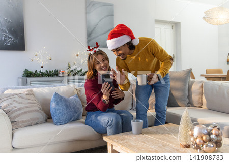 Couple wearing Santa hat and reindeer headband sitting in living room on couch, viewing smartphone 141908783
