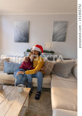 Couple cuddling on beige sectional sofa at home, wearing Santa hat beside coffee table with mugs 141908792