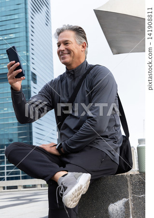 Middle-aged man sitting on concrete ledge at urban plaza wearing dark activewear holding smartphone Middle-aged man sitting on concrete ledge at urban plaza wearing dark activewear holding smartphone 141908811