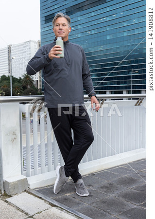 Middle-aged man in athletic wear leaning on bridge railing, drinking from water bottle with earbud Middle-aged man in athletic wear leaning on bridge railing, drinking from water bottle with earbud 141908812