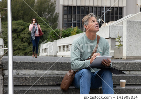 Middle-aged man sitting on steps at urban plaza sketching in sketchbook with coffee, messenger bag Middle-aged man sitting on steps at urban plaza sketching in sketchbook with coffee, messenger bag 141908814