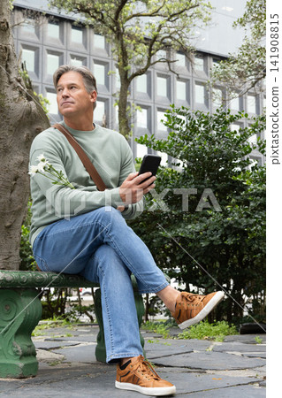 Middle-aged man sitting on green bench in urban plaza holding phone and white flowers in sweatshirt Middle-aged man sitting on green bench in urban plaza holding phone and white flowers in sweatshirt 141908815