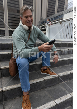 Senior man sitting on concrete steps checking smartphone while wearing headphones and crossbody bag 141908819