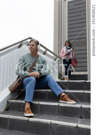Commuters sitting on steps by office tower, man wearing headset using tablet, woman holding cup 141908821
