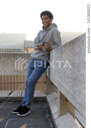 Teenage male leaning on concrete railing on rooftop at sunset smiling while wearing hoodie, watch Teenage male leaning on concrete railing on rooftop at sunset smiling while wearing hoodie, watch 141908822