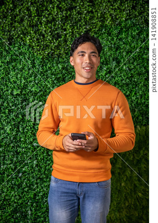 Adult Asian man standing in front of green hedge wall holding phone wearing orange sweatshirt jeans 141908851