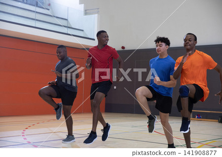 Diverse male team performing high-knee drills in school gym on hardwood court, knee brace visible Diverse male team performing high-knee drills in school gym on hardwood court, knee brace visible 141908877