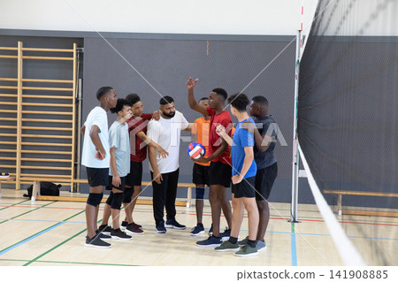 Diverse male volleyball team huddling near net post on gym court holding blue-white-red volleyball Diverse male volleyball team huddling near net post on gym court holding blue-white-red volleyball 141908885