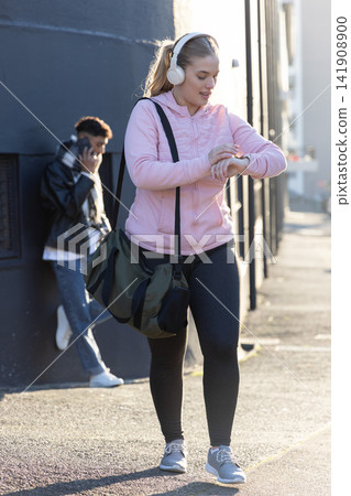 Woman walking on sunny sidewalk in pink hoodie, leggings, checking smartwatch carrying green duffel Woman walking on sunny sidewalk in pink hoodie, leggings, checking smartwatch carrying green duffel 141908900