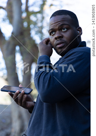 African American man standing among trees in navy pullover holding phone adjusting earbud African American man standing among trees in navy pullover holding phone adjusting earbud 141908905