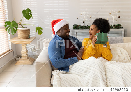African American couple exchanging green gift laughing on couch with Santa hat mustard sweater African American couple exchanging green gift laughing on couch with Santa hat mustard sweater 141908910