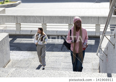 Diverse female students walking up campus stairs, carrying backpack, notebooks, coffee, copy space Diverse female students walking up campus stairs, carrying backpack, notebooks, coffee, copy space 141908911