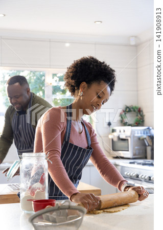 African American couple baking at home in striped aprons, woman rolling dough with wooden pin African American couple baking at home in striped aprons, woman rolling dough with wooden pin 141908913