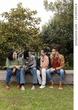 Diverse friends sitting on bench in park chatting, holding phone, wearing scarves and jeans Diverse friends sitting on bench in park chatting, holding phone, wearing scarves and jeans 141908914