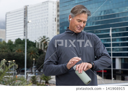 Middle aged man pausing on urban walk wearing athletic top holding green bottle checking smartwatch 141908915