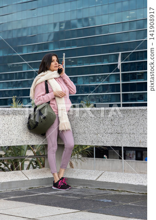 Female leaning on balustrade on plaza in pink hoodie, speaking on smartphone, holding olive duffel Female leaning on balustrade on plaza in pink hoodie, speaking on smartphone, holding olive duffel 141908917