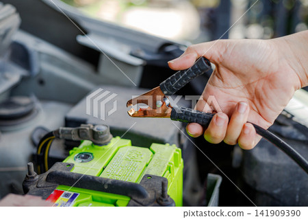 Hand holding jumper cable clamp near car battery Hand holding jumper cable clamp near car battery 141909390