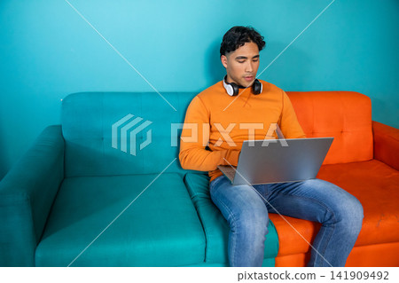 Man sitting on two-tone sofa wearing orange sweatshirt with headphones using laptop, copy space 141909492