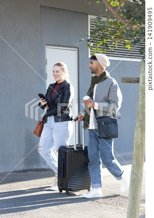 Couple walking beside grey facade, woman holding phone, man pulling black suitcase, sipping coffee Couple walking beside grey facade, woman holding phone, man pulling black suitcase, sipping coffee 141909495