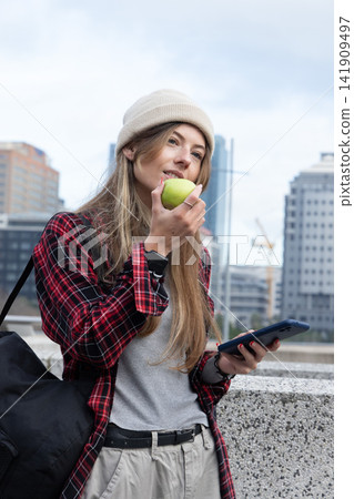 Female commuter wearing cream beanie leaning on concrete railing holding green apple and phone Female commuter wearing cream beanie leaning on concrete railing holding green apple and phone 141909497