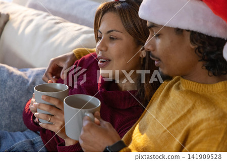 Couple sitting close on sofa at home, holding gray mugs and wearing Santa hat, ring visible Couple sitting close on sofa at home, holding gray mugs and wearing Santa hat, ring visible 141909528
