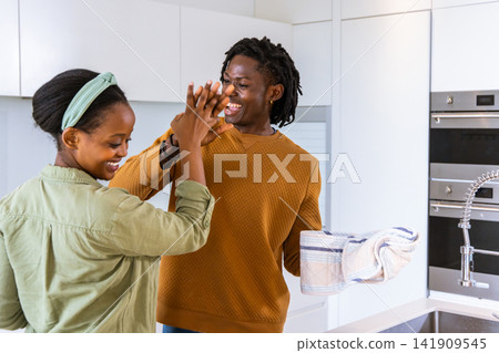 African American couple high-fiving at kitchen sink woman mint shirt man rust sweater holding towel African American couple high-fiving at kitchen sink woman mint shirt man rust sweater holding towel 141909545
