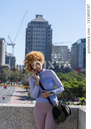 African American woman leaning on stone railing talking on phone, checking watch with bag in city 141909597