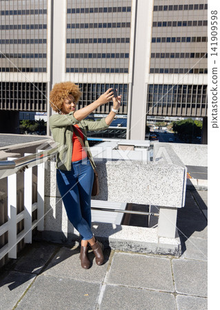 African American woman leaning on parapet holding smartphone for selfie on rooftop near handrail 141909598