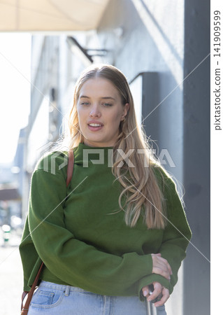 Woman walking on city sidewalk beside dark wall, wearing green sweater and holding metal handle 141909599
