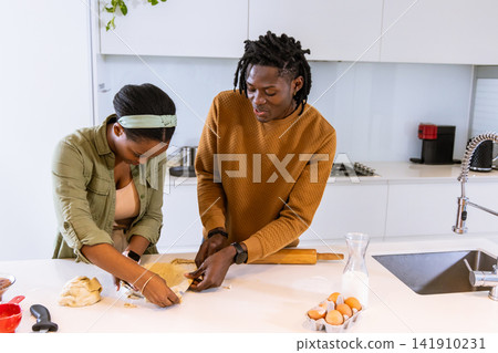 African American couple in casual wear prepping dough with rolling pin and eggs at kitchen island 141910231