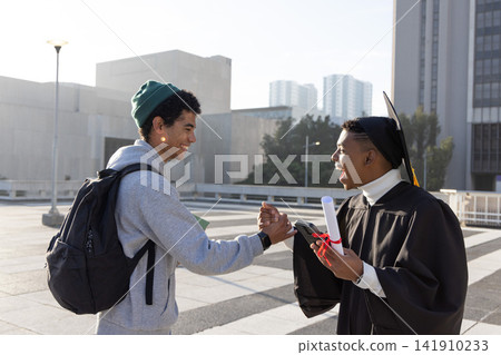 Diverse male friends cheering on plaza, graduate wearing gown holding diploma and phone, copy space Diverse male friends cheering on plaza, graduate wearing gown holding diploma and phone, copy space 141910233