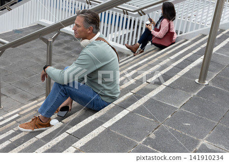 Diverse pair sitting on public steps at stairwell, man wearing headphones, both holding smartphones 141910234