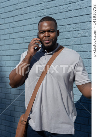 African American man 30s standing, talking on phone against blue brick wall with gray polo, satchel African American man 30s standing, talking on phone against blue brick wall with gray polo, satchel 141910758