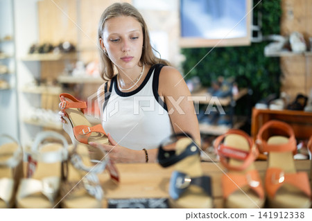 Girl spends time in store and pick up summer sandals, shopping in shoe boutique. Female buyer examines product, turns shoe sample in hands and makes purchase decision. Girl spends time in store and pick up summer sandals, shopping in shoe boutique. Female buyer examines product, turns shoe sample in hands and makes purchase decision. 141912338