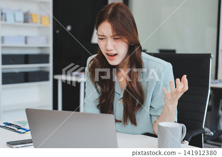 Asian businesswoman having a stressful phone call, holding a pen, and looking troubled while working on her laptop with financial documents and a calculator on her office desk Asian businesswoman having a stressful phone call, holding a pen, and looking troubled while working on her laptop with financial documents and a calculator on her office desk 141912380