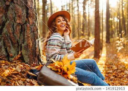 Pretty young woman in hat reading interesting book near tree in autumn forest. Enjoying nature Relax Pretty young woman in hat reading interesting book near tree in autumn forest. Enjoying nature Relax 141912913