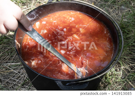 Delicious traditional Ukrainian red borscht soup cooking in a cast iron pot outdoors on fire, stirred with a ladle, showcasing fresh vegetables and rich broth for camping and nature travel food. Delicious traditional Ukrainian red borscht soup cooking in a cast iron pot outdoors on fire, stirred with a ladle, showcasing fresh vegetables and rich broth for camping and nature travel food. 141913085