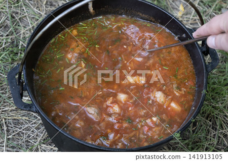 Delicious traditional Ukrainian red borscht soup cooking in a cast iron pot outdoors on fire, stirred with a ladle, showcasing fresh vegetables and rich broth for camping and nature travel food. Delicious traditional Ukrainian red borscht soup cooking in a cast iron pot outdoors on fire, stirred with a ladle, showcasing fresh vegetables and rich broth for camping and nature travel food. 141913105