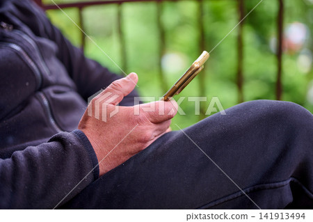 A person sits outdoors and looks at their smartphone. The scene has green plants in the background. It is a relaxed setting during daytime A person sits outdoors and looks at their smartphone. The scene has green plants in the background. It is a relaxed setting during daytime 141913494