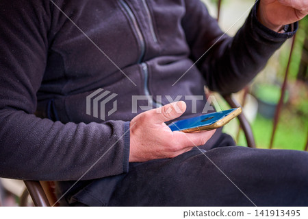 A person sits on a chair outside, holding a smartphone. The background shows green plants and a relaxed outdoor space. It is daytime and the weather looks nice A person sits on a chair outside, holding a smartphone. The background shows green plants and a relaxed outdoor space. It is daytime and the weather looks nice 141913495