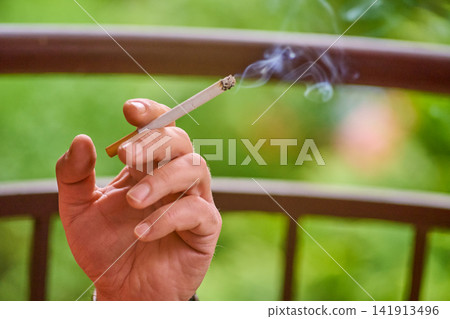 A person holds a cigarette in their hand while smoke rises into the air. This scene takes place in an outdoor area surrounded by greenery A person holds a cigarette in their hand while smoke rises into the air. This scene takes place in an outdoor area surrounded by greenery 141913496